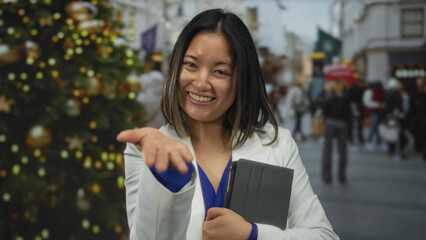 Woman smiling outdoors in a city street holding a tablet with festive decorations and reaching out with a friendly gesture.