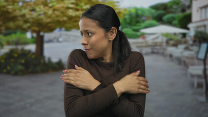 Woman shivering outdoors in a street restaurant terrace on a cool autumn day, expressing cold and discomfort.