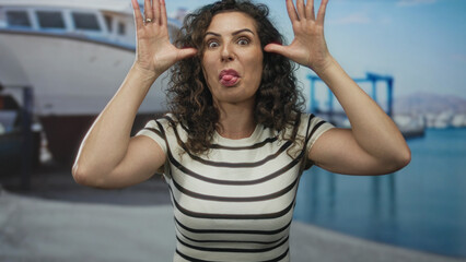 Woman sticks out her tongue and spreads her hands by her eyes at street with a boat docked behind...