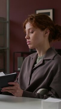 Serious young Caucasian buinesswoman sitting at deskin modern office, using latop and taking notes in noebook