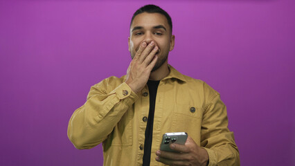 Young hispanic man covers mouth with hand while holding smartphone in purple studio setting;...