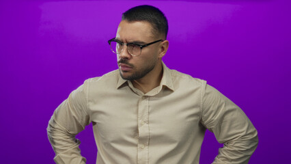 Young hispanic man wearing glasses pointing finger forward in vibrant purple studio; determination focus authority.