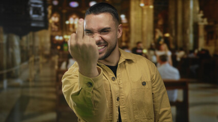 Young hispanic man flips middle finger and clenches fists inside ornate church building with wooden pews and chandeliers; defiance rebellion resistance insult.