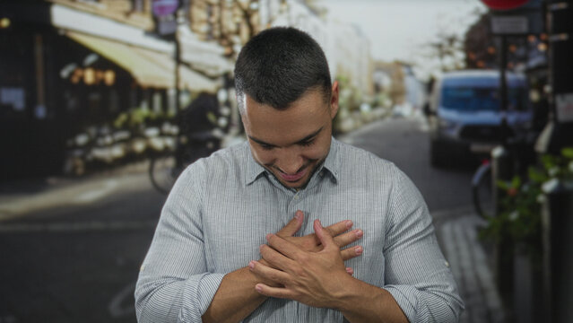 Smiling hispanic man in striped shirt crosses hands over chest on sunlit street; gratitude sincerity.