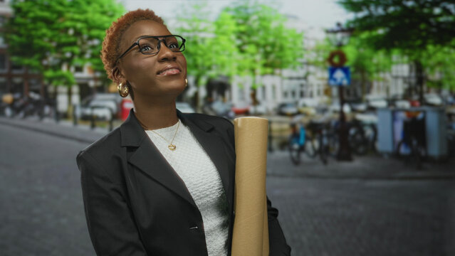 Woman in black blazer holds blueprints under green foliage on urban street by building facade; ambition.