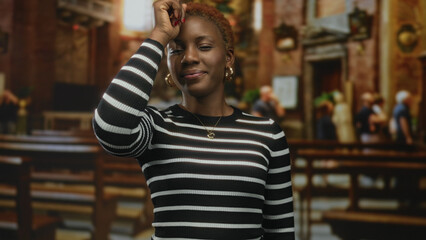 Smiling young woman stands upright and gracefully touches her forehead in historic building;...