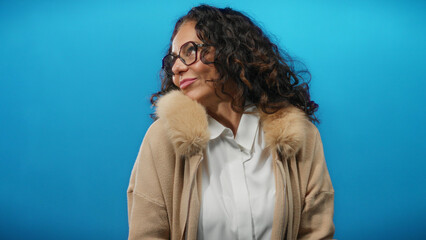 Woman with curly hair wearing glasses and a beige coat poses against a vibrant blue background, exuding elegance and confidence in an isolated studio setting.
