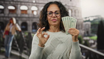 Woman holding american dollars near the roman coliseum makes an ok gesture, combining elements of travel and finance in an iconic italian outdoor city setting.