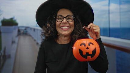 Woman wearing witch hat covers face with hand while holding pumpkin basket on boat deck; joy surprise festivity celebration.