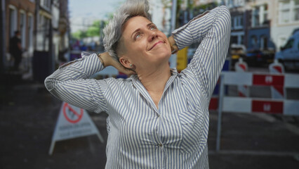 Woman touches neck on building site street lined with parked vans and barricades in daylight;...