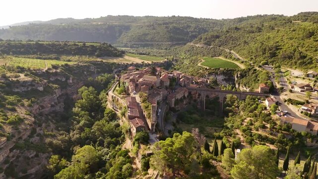 Aerial view of Minerve, a village perched on a cliff, linked by an ancient stone bridge, surrounded by lush green forests and vineyards, Minerve, Occitanie, France.