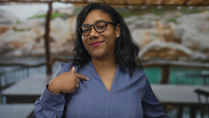 Woman in blue blouse wearing glasses points finger to chest on street during daytime; self confidence.