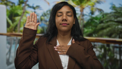 African american woman in brown blazer raises hand and places hand on chest in park scene; honor...