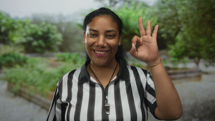 Woman referee in striped shirt makes ok sign with hand while holding whistle in park; confidence.