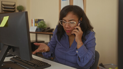 African american woman holds phone to ear with furrowed brow while looking at monitor in office;...