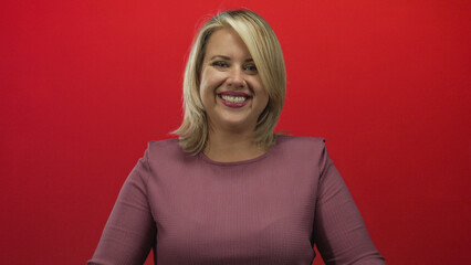 Obraz premium Young blonde woman smiles broadly and extends arms with hands against red studio wall; invitation joy.