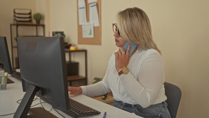 Young blonde woman writes with pen at desk in building beside computer while holding phone to ear;...