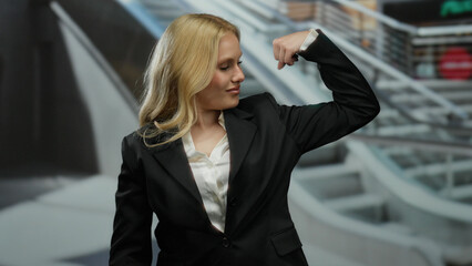Young blonde woman in a mall flexing arm power in business attire, showcasing confidence and success against an urban shopping center backdrop with escalators.