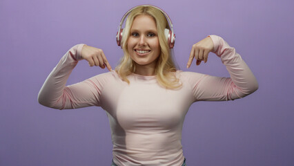 Fototapeta premium Young blonde woman wearing headphones against a purple background smiling and pointing downwards with both hands in a playful manner
