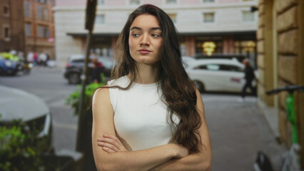 Fototapeta premium Young hispanic woman in a white tank top with crossed arms stands on a crowded city street with parked cars; defiance.