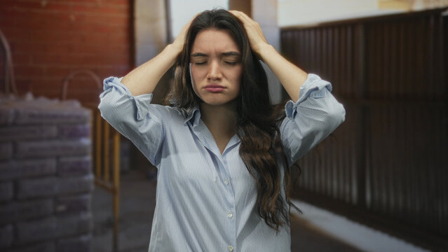 Young woman holds head with both hands for headache in building under construction with closed eyes; stress.