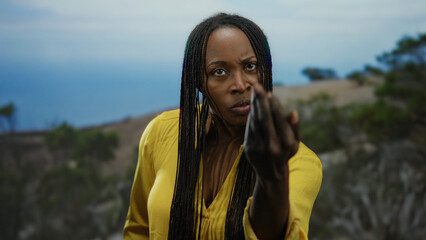 Woman with braids wearing yellow shirt at seaside pointing with intense expression against ocean...