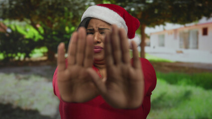 Woman in santa hat and red sweater with fearful expression in a park, gesturing fear with hands,...
