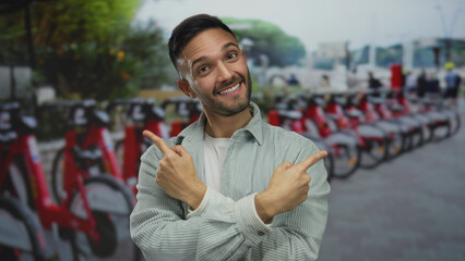 Young man smiling and pointing his fingers in an urban street with rows of bicycles creating a...