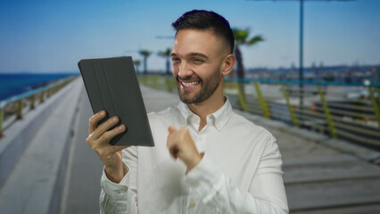 Man smiling holding tablet while on video call at sunny seaside promenade with palm trees and ocean...