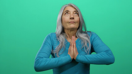 Senior woman with grey hair in prayer pose against a bright green background, wearing a blue...