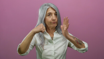 Senior woman with grey hair performs gesture with raised fists against isolated pink background in...