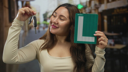 Hispanic woman smiling holds dangling car keys and green learner plate outside restaurant building...