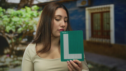 Young hispanic woman holds green learner sign beside building outdoors under diffuse daylight; driving ambition.