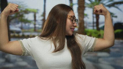 Young hispanic woman flexes bare arms and clenches fists in sunlit green park with palm trees...