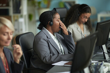 Black woman wearing headset yawning while working at computer in office, sitting next to Caucasian...