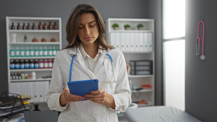 Woman doctor in clinic using tablet while standing in modern hospital room with medical equipment and shelves in background.