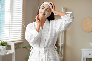 Beautiful young happy woman in bathrobe with applied clay mask sitting at home