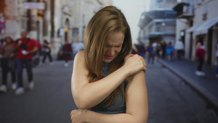 Woman in blue tank top holding shoulder in busy urban street amid blurred crowd; physical...