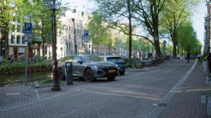 Blurred view of amsterdam street with parked cars, canal, trees, and architecture in the background on a sunny day.
