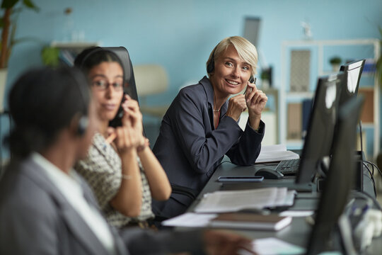 Caucasian middle aged woman smiling while wearing headset and sitting at desk with multiethnic young adult colleagues working in modern office call center environment - Powered by Adobe