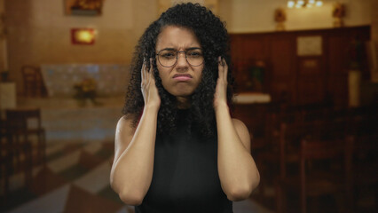 Young hispanic woman furrows brow and adjusts round metal glasses inside a building as she...