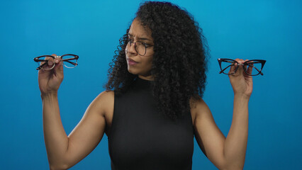 Woman choosing between glasses against blue background expressing contemplation and uncertainty with two different eyewear options showcasing style and decision making