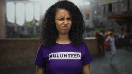 Hispanic woman in volunteer shirt stands outdoors on historic town street with thoughtful...
