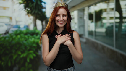 Woman with red hair and crown smiling gracefully, standing outdoors on a city street with hands on chest, exuding beauty and elegance.
