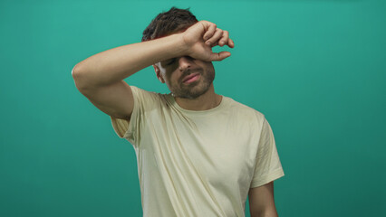 Young hispanic man raising arm to forehead gesture in white tshirt in studio against teal walls;...