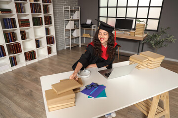 Female graduating student sitting at table with stack of books and laptop in library