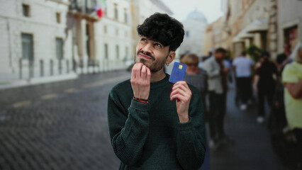 Young man holding creditcard on urban street pondering financial decision considering people and...