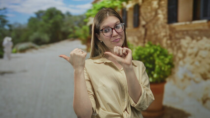 Fototapeta premium Blonde woman wearing glasses points thumbs to herself in front of ancient stone building; confidence.