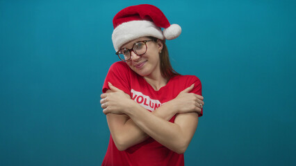 Woman extends arms in open hug gesture wearing volunteer shirt and santa hat in blue studio; welcoming warmth.