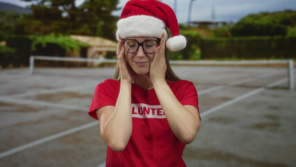 Obraz premium Young volunteer woman wearing glasses and santa hat touches face and smiles on tennis court outdoors; serenity.